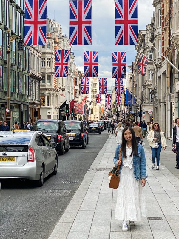 Lady in white midi skirt and shirt with a light blue demin jacket, walking elegantly along the famous New Bond Street in London. The street is decorated with Union Jacks as part of the Platinum Jubilee display. Demonstrating that dressing the part an important part in how to speak elegantly.