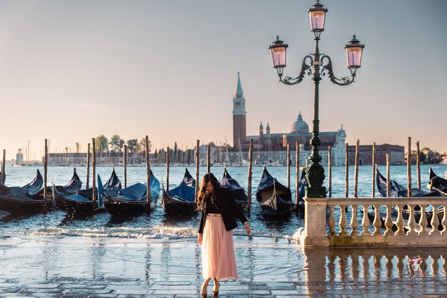 How to be an Elegant woman: Finally Seen, Heard, and Command Respect 16 An elegant woman dressed in a flowing pastel skirt and black jacket stands near a row of gondolas docked on a Venetian canal. The setting features a scenic view of San Giorgio Maggiore in the background, with its iconic bell tower and dome, under a clear blue sky. A classic wrought-iron lamppost adds charm to the luxurious travel experience in Venice.