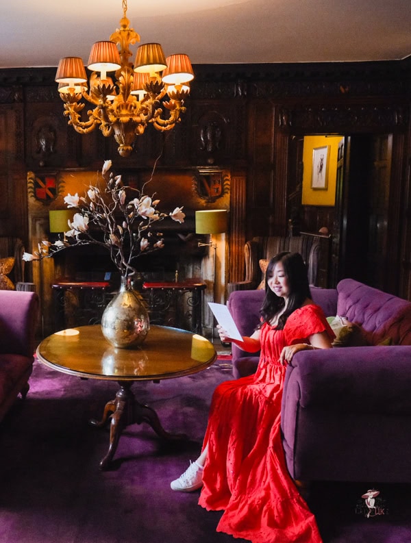 How to be an Elegant woman: Finally Seen, Heard, and Command Respect 15 A woman in an elegant red dress sits on a plush purple sofa in a luxurious, wood-panelled room. She holds a piece of paper while surrounded by a warm ambience, including a golden vase with magnolia flowers on a polished round table and an ornate chandelier overhead. The setting exudes sophistication and charm, perfectly complementing her graceful presence.