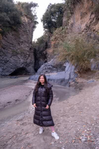 Exclusive trips to Mount Etna from catania: Elegant and Stress-free Options 32 A woman in a black puffer coat standing on Alcantara's Gorge valley with a clear view of the mountainous rock formations from the hot lava flow created these interesting, unique ripples in the rocks.